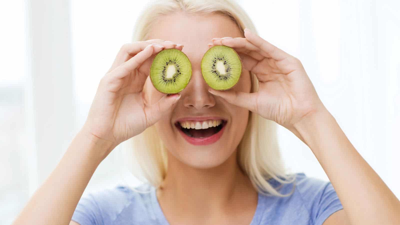 A person with long blonde hair is smiling and playfully holding two halves of a kiwi fruit in front of their eyes. They are wearing a light blue shirt and appear to be indoors with a bright background.