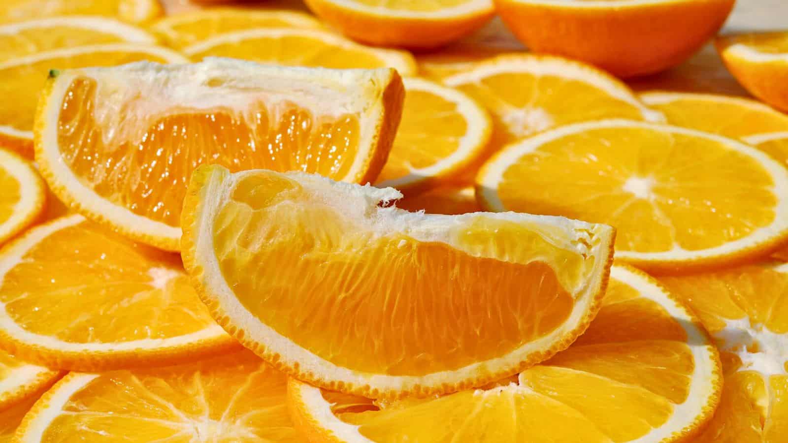 A close-up of many sliced oranges. In the foreground, there are two orange wedges placed on top of circular orange slices. The vibrant, juicy texture of the fruit is visible, with the outer peel framing the bright orange interior.