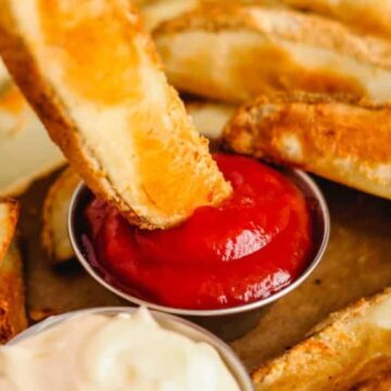 Close-up of crispy potato wedges being dipped into a small container of red ketchup, with a separate dish of white dipping sauce nearby. These carb-loving delights are golden brown, suggesting they are freshly cooked.