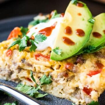 A close-up of a slice of casserole served on a gray plate. The dish includes ground meat, melted cheese, and red bell pepper, topped with sliced avocado, sour cream, a drizzle of hot sauce, and garnished with fresh cilantro. A fork is placed beside it.
