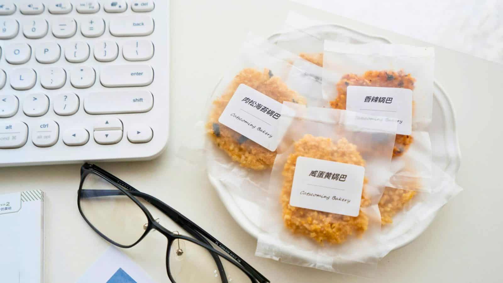 A plate with four packaged cookies sits on a white surface. Each package has a label with Chinese characters and the text "Chainspring Bakery." Nearby are a white keyboard, a pair of black-rimmed glasses, and part of a notebook.