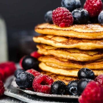 A stack of pancakes topped with raspberries and blueberries graces a plate surrounded by more berries. In the background, a small glass bottle and an auto draft container with syrup are partially visible.