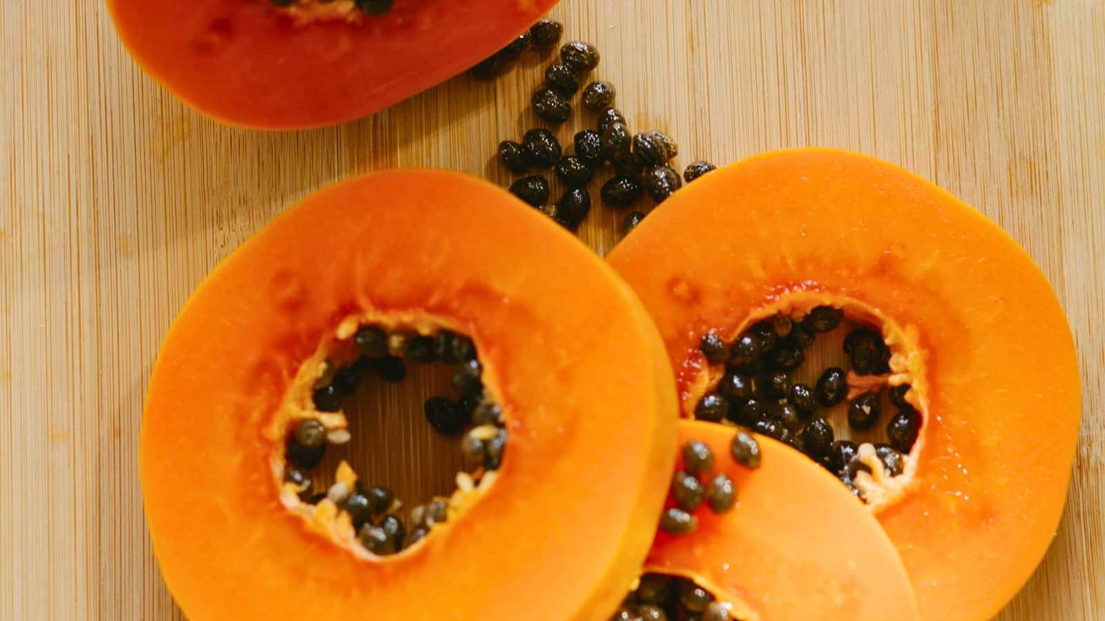 Slices of a ripe papaya with black seeds are displayed on a wooden cutting board. The bright orange flesh contrasts with the scattered seeds.