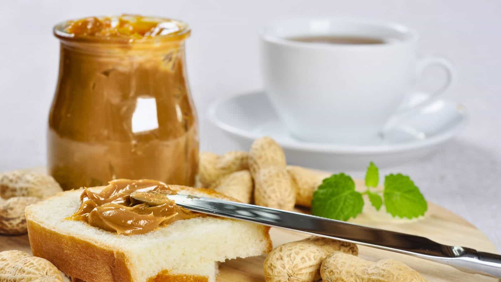 A knife spreads peanut butter on a slice of bread, surrounded by peanuts. A jar of peanut butter is open nearby. A white cup of coffee sits in the background on a white saucer. A sprig of green leaves is also visible.