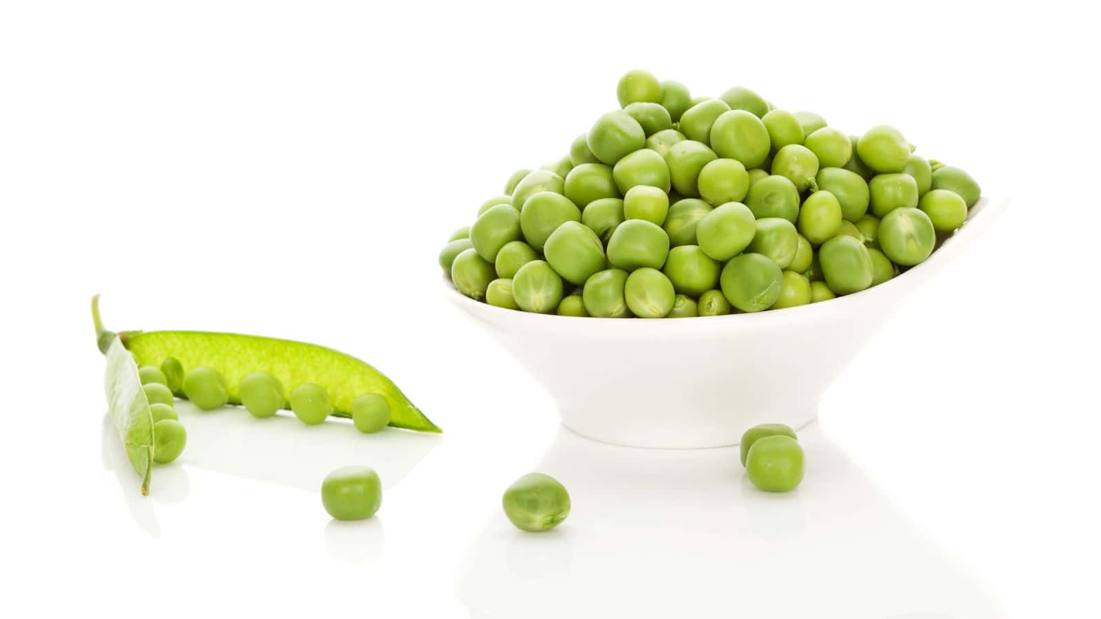 A white bowl filled with fresh green peas is placed on a reflective surface. Beside it, an open pea pod with several peas still inside is positioned alongside a few loose peas. The background is a bright white.