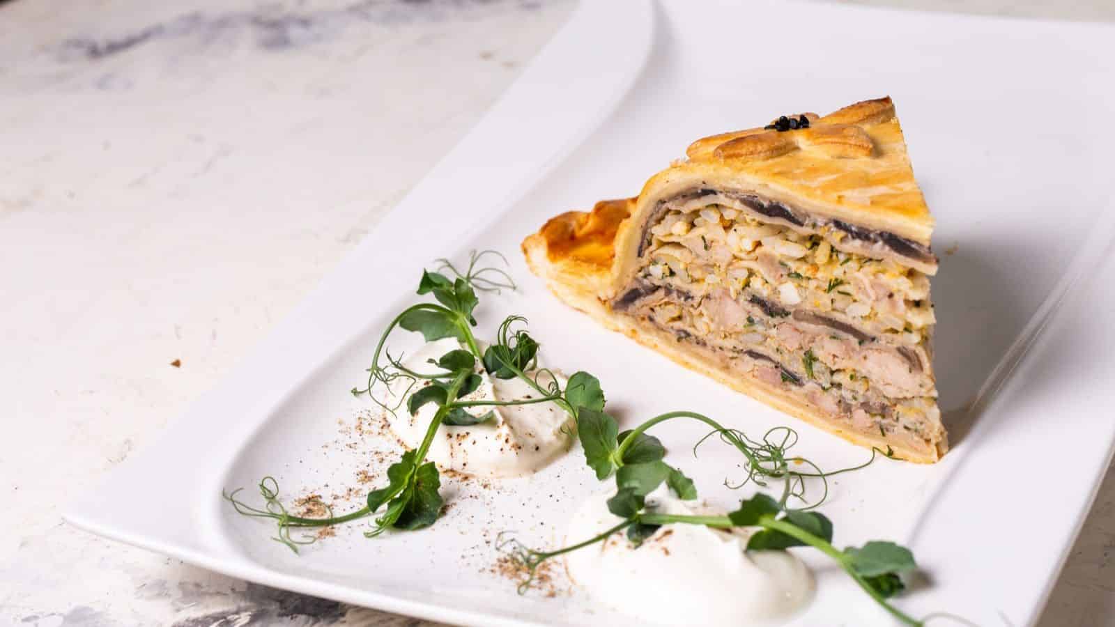 A slice of layered savory pie with mushrooms and meat on a white plate, garnished with two dollops of cream and topped with pea shoots, with sprinkles of seasoning nearby.
