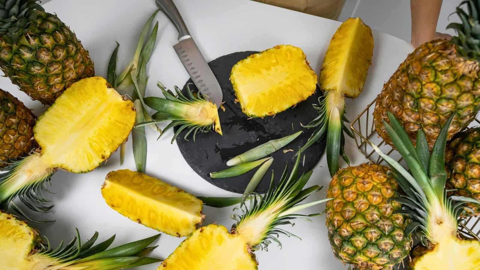 A variety of pineapples are sliced open and displayed on a white surface. A large knife rests on a black cutting board at the center, surrounded by pineapple pieces and leaves. A hand is visible holding a whole pineapple on the right.