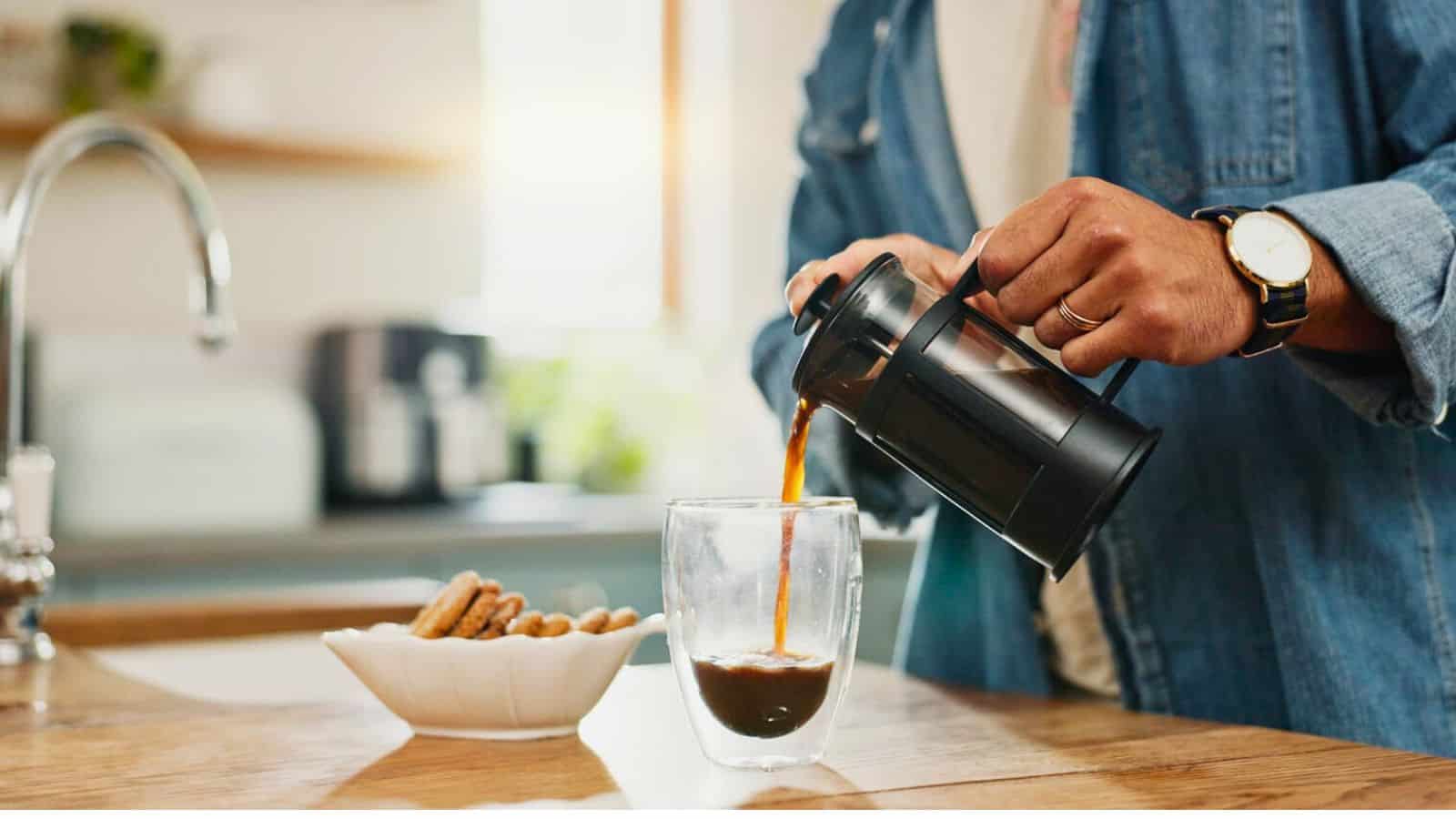 A person pours coffee from a French press into a glass on a wooden kitchen countertop. A white bowl filled with cookies is nearby. Sunlight streams through a window, illuminating the scene.