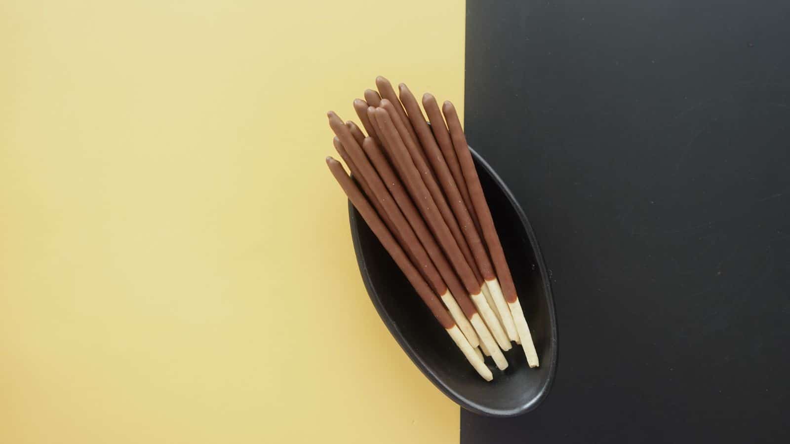 A black oval dish holds a neatly arranged stack of chocolate-covered biscuit sticks, an international snack delight. The dish sits on a background split between yellow on the left and dark gray on the right, offering a quirky contrast that might intrigue adventurous Americans.