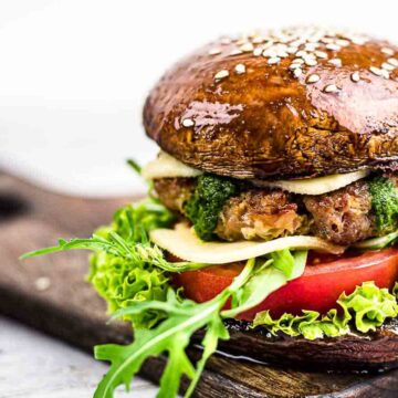 A keto burger with a portobello mushroom bun, topped with fresh greens, juicy tomato, melted cheese, and savory pesto sauce, all artfully presented on a rustic wooden board.