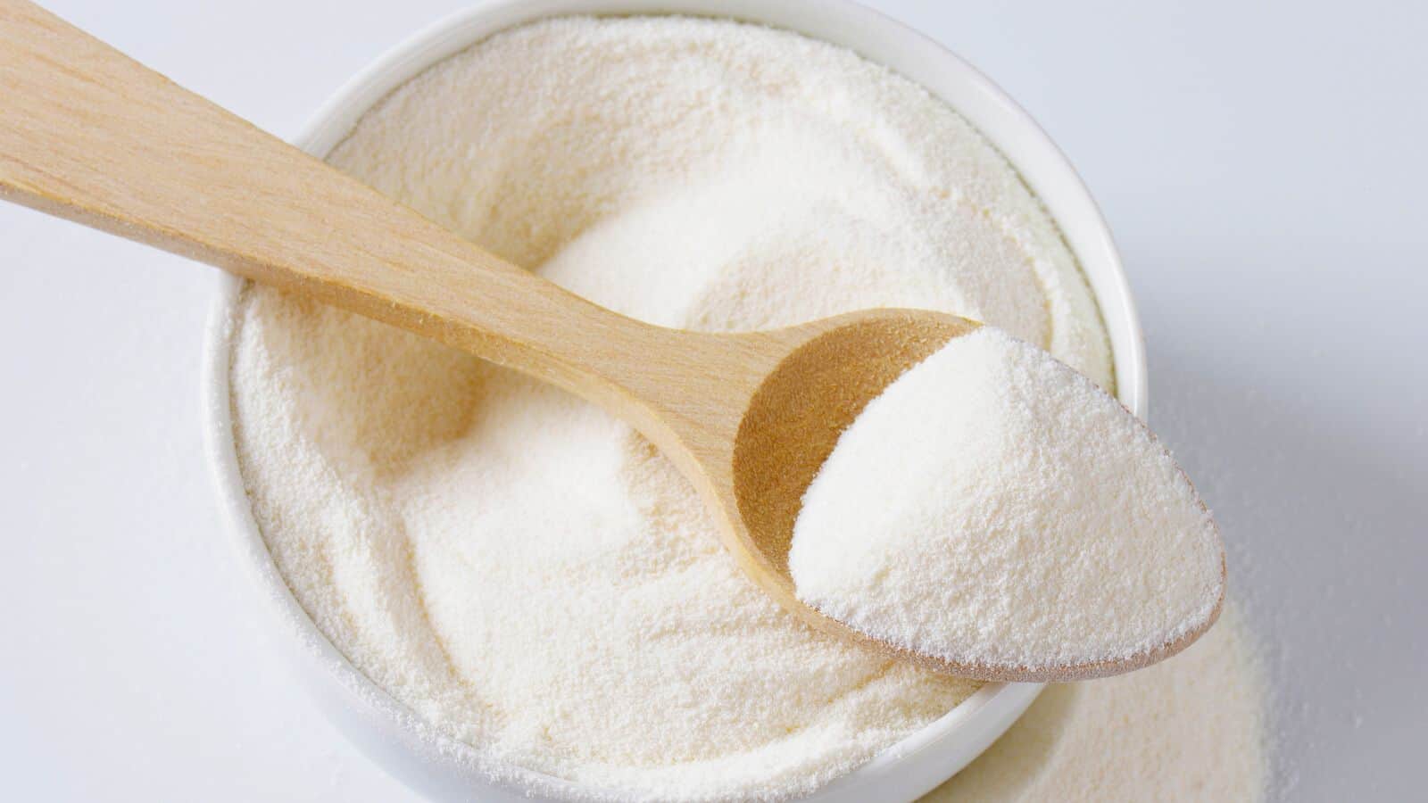 A wooden spoon rests on a bowl filled with white powdered milk. The spoon is slightly buried in the powder, showing a small amount of the product on its surface. The background is a clean, light surface.