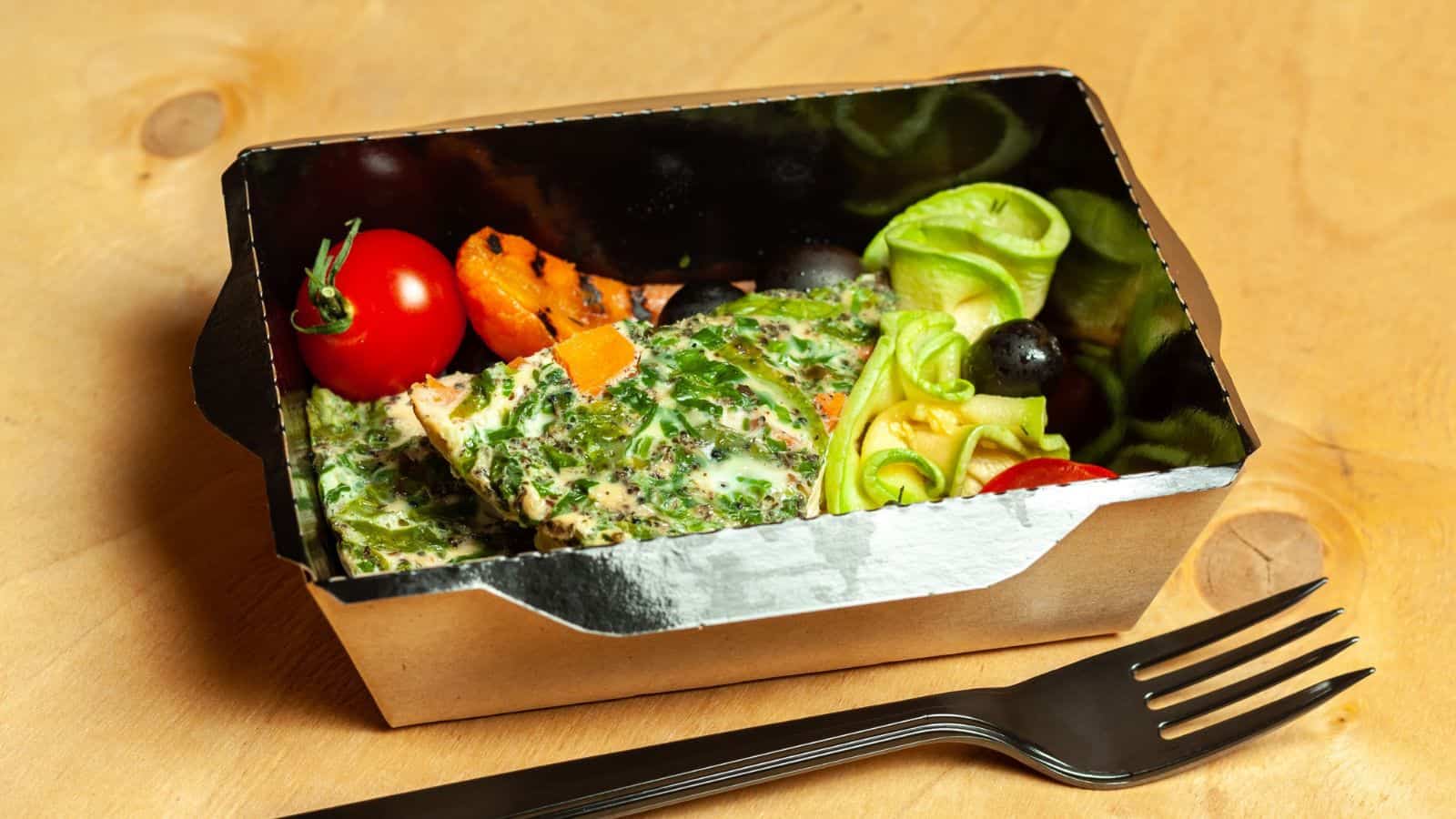 A takeout container on a wooden table holds a meal with a grilled chicken breast topped with herbs, a side of lettuce, cherry tomatoes, a slice of carrot, grilled vegetables, black olives, and a fork placed beside the container.