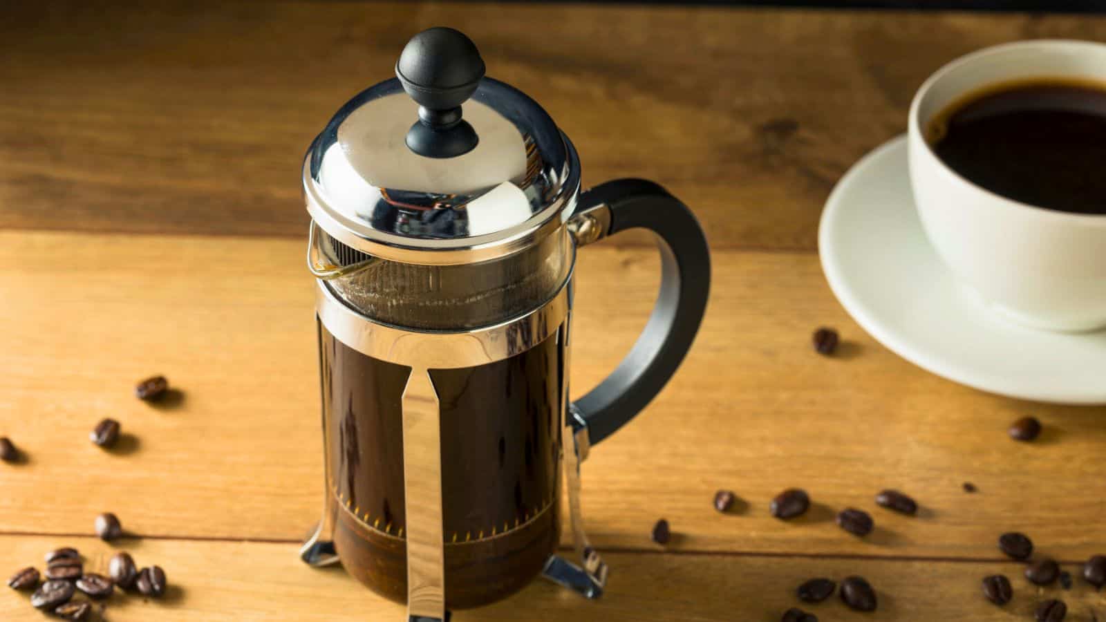 A French press filled with coffee on a wooden table, surrounded by scattered coffee beans. To the right, there is a white cup and saucer with coffee.