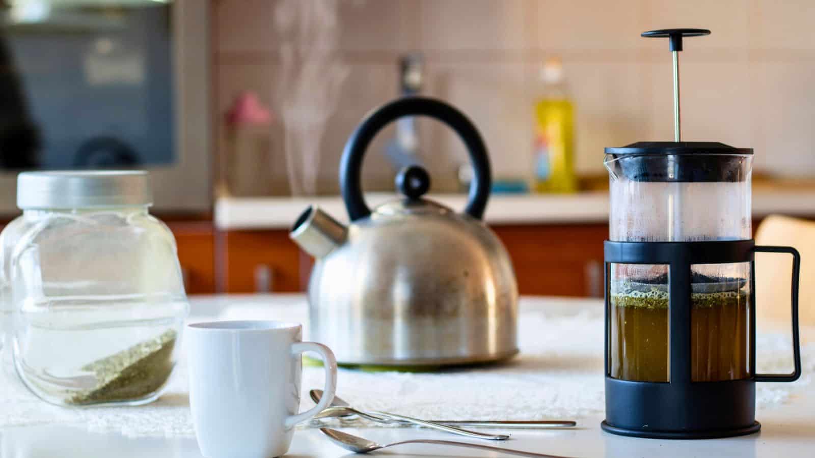A kitchen table with a French press containing brewed coffee, a white mug, two teaspoons, a glass jar with a white substance, and a metal kettle in the background.