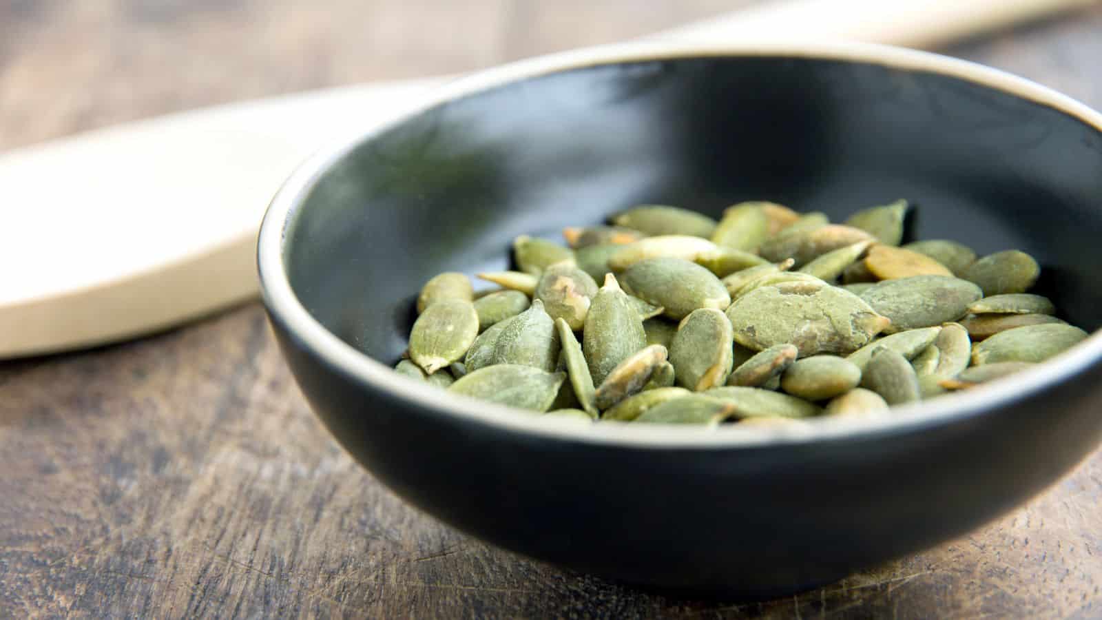 A black bowl filled with green pumpkin seeds sits on a wooden surface. A wooden spoon is partially visible in the background.