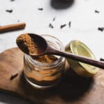A wooden spoon with ground pumpkin pie spice rests on a glass jar filled with the same blend. The jar is on a wooden board with its lid nearby, while a few scattered cloves and a cinnamon stick linger in the background, ready to infuse your 4-ingredient pumpkin pie with warmth.
