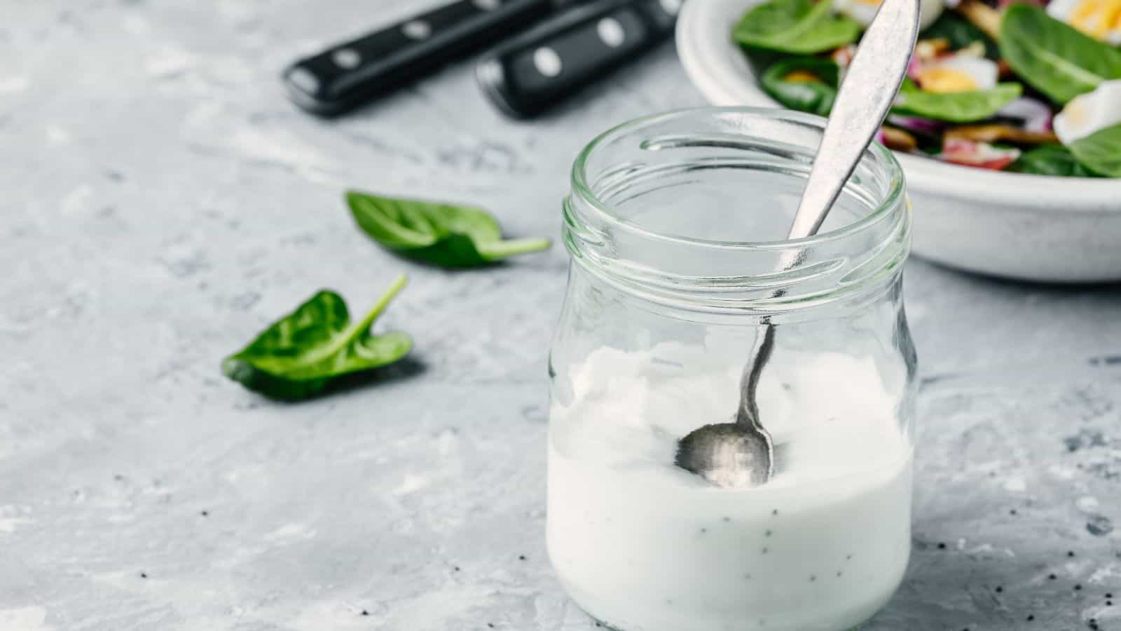 A glass jar of creamy dressing with a spoon inside is placed on a textured gray surface. In the background, a bowl of mixed salad greens is partially visible, with a few spinach leaves scattered around. Three black-handled utensils are nearby.