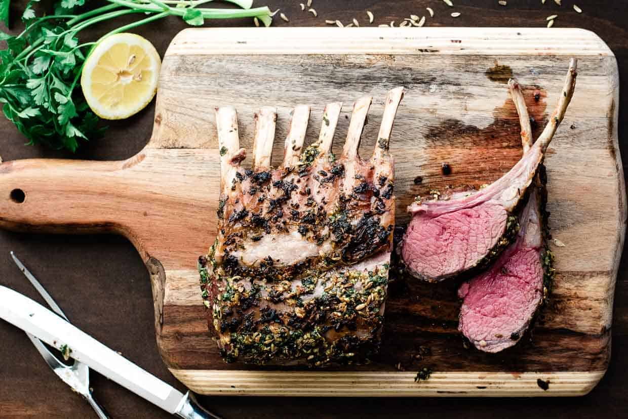 A cooked rack of lamb rests on a wooden cutting board, embodying the spirit of Thanksgiving without turkey. Two slices are cut beside the herb-seasoned meat. A lemon half and parsley garnish the scene, accompanied by a carving knife and fork, offering a twist on traditional feastings.