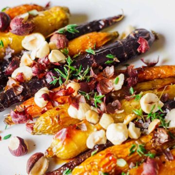 A dish of roasted carrots with hazelnuts in shades of orange and purple is garnished with thyme leaves and edible flower petals. The presentation is on a white plate.