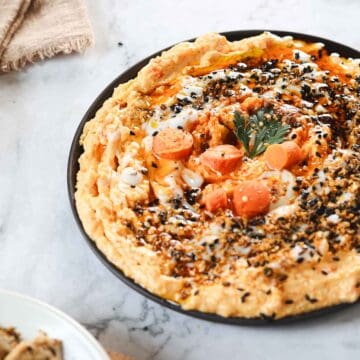 A plate of creamy hummus topped with chopped carrots, parsley, and black sesame seeds. The dish is garnished with a drizzle of oil. A partial view of a cloth napkin and a bowl with slices of bread can be seen nearby on a marble surface.
