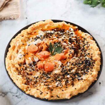 A plate of hummus topped with sliced carrots, a sprig of parsley, and sprinkled with black sesame seeds and spices. The dish is set on a marble surface, with a piece of burlap and some greens partially visible in the background.