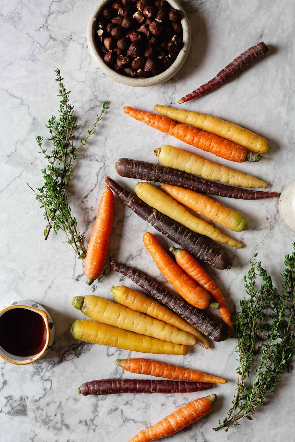 A variety of multicolored carrots are arranged on a marble surface alongside fresh thyme sprigs. A bowl of nuts and a small cup with a dark liquid are also present.