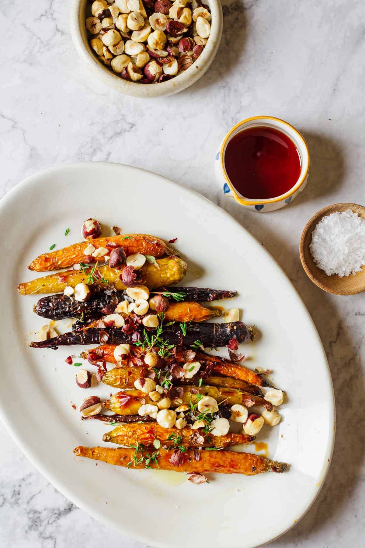 A plate of roasted carrots topped with chopped hazelnuts and herbs is set on a marble surface. Nearby are a small bowl of hazelnuts, a cup of red liquid, and a wooden bowl of sea salt.