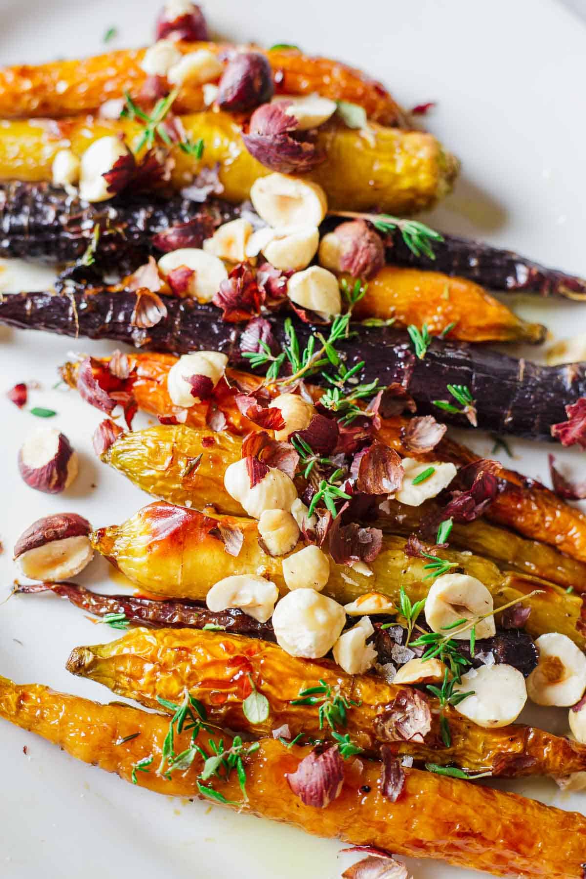 A plate of roasted heirloom carrots topped with hazelnuts and fresh thyme. The carrots are arranged in a colorful assortment of orange, yellow, and purple. The hazelnuts add texture, while the thyme provides a garnish.