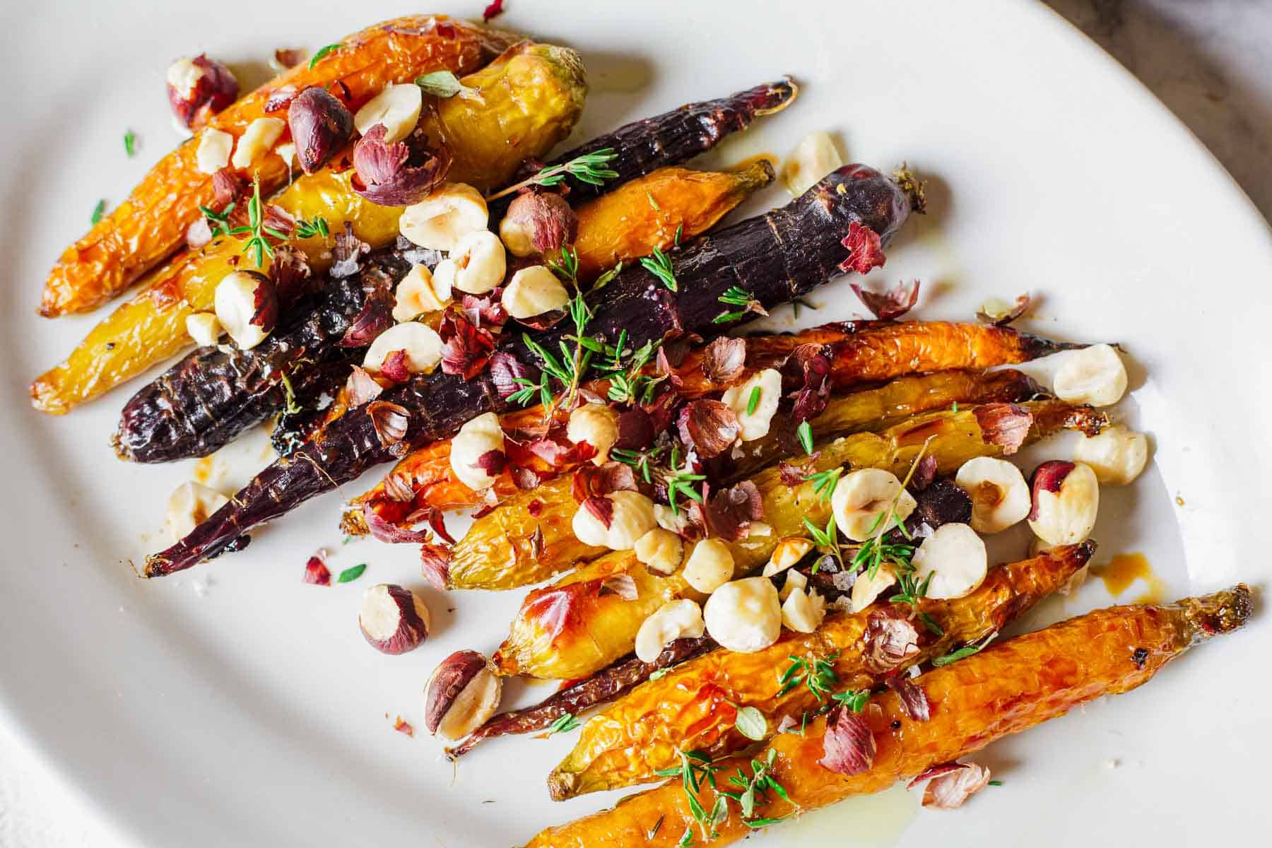 A plate of roasted heirloom carrots, garnished with chopped hazelnuts and fresh thyme. The carrots are in shades of orange and purple, drizzled with a light glaze.