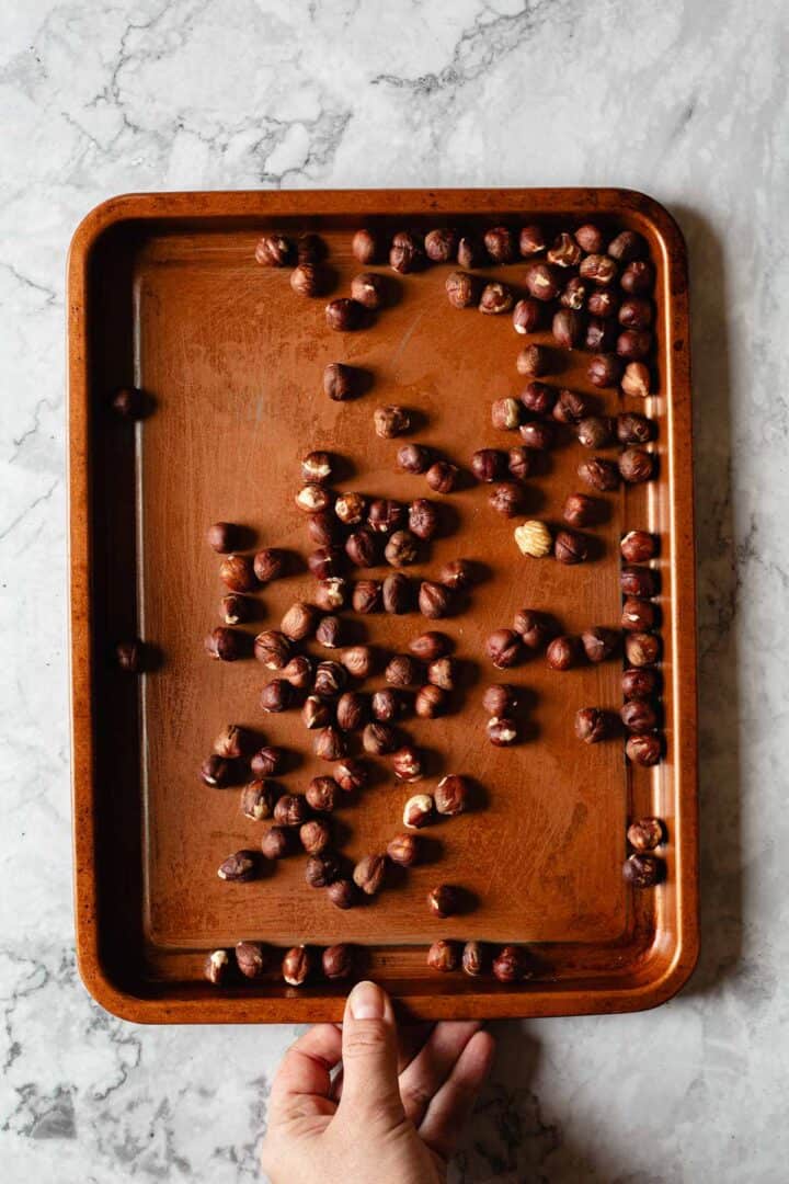 A hand holds a copper baking tray scattered with roasted hazelnuts on a marble countertop.