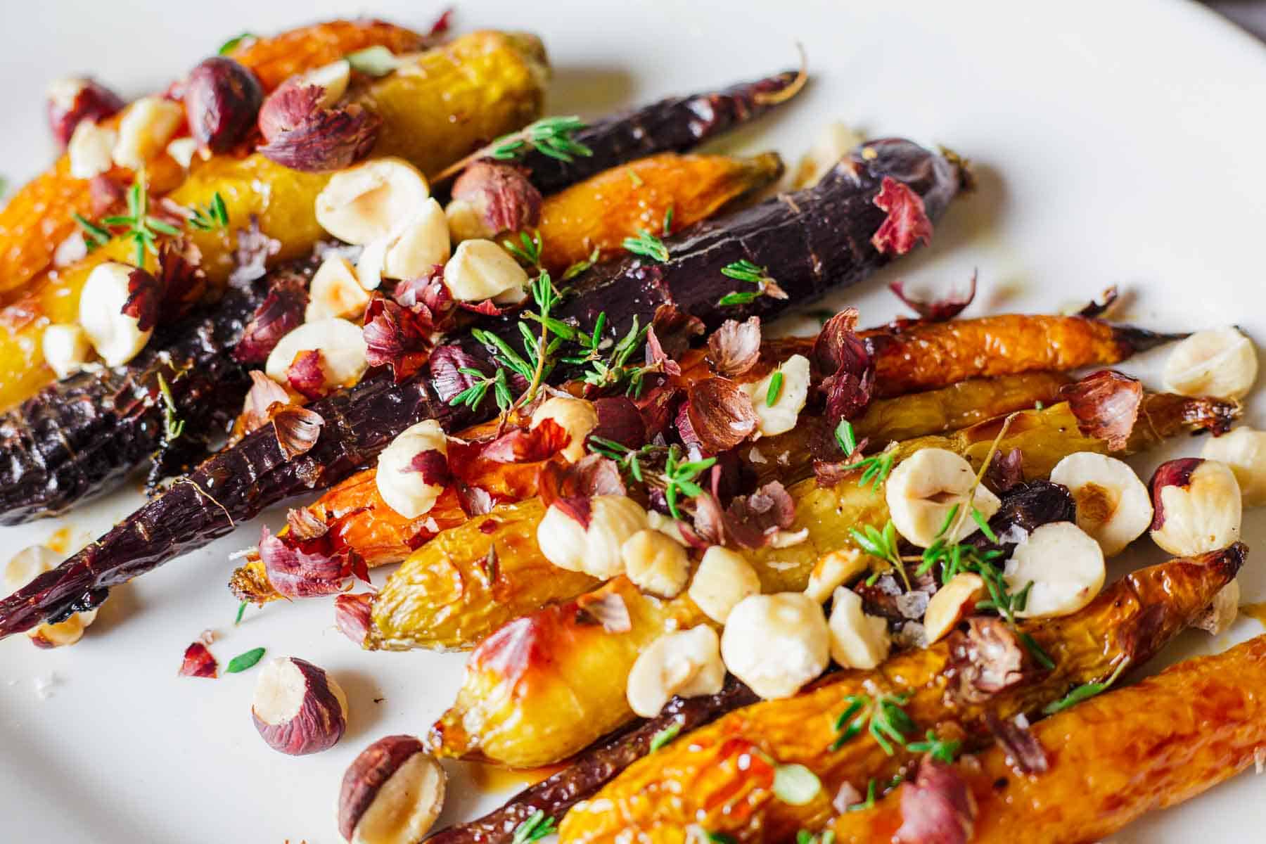 A plate of roasted carrots in shades of orange and purple, garnished with hazelnuts, fresh herbs, and salt flakes. The carrots have a glossy texture, suggesting they are seasoned and roasted, providing a colorful and vibrant presentation.