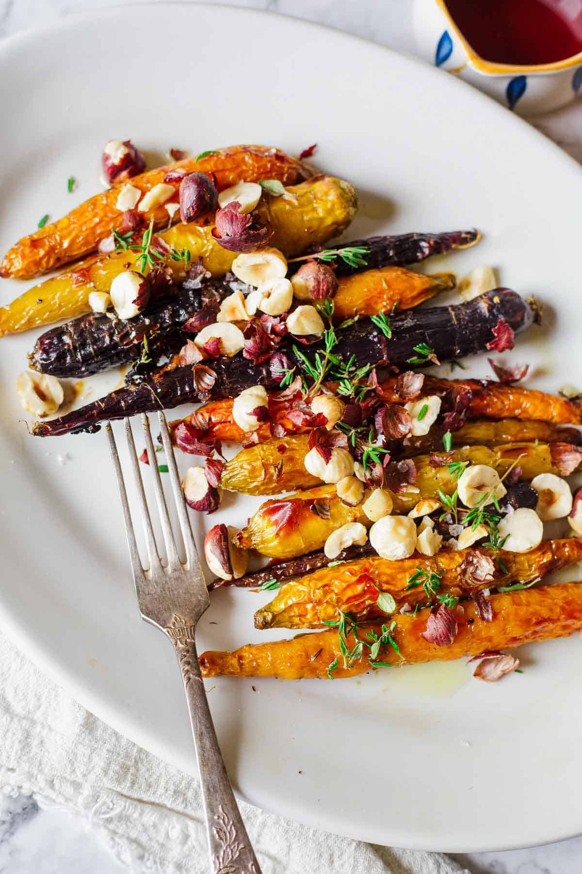 A plate of roasted mixed-color carrots topped with chopped nuts and herbs. A fork rests on the plate, and a white napkin is partially visible underneath. A small cup with a red liquid is in the background.