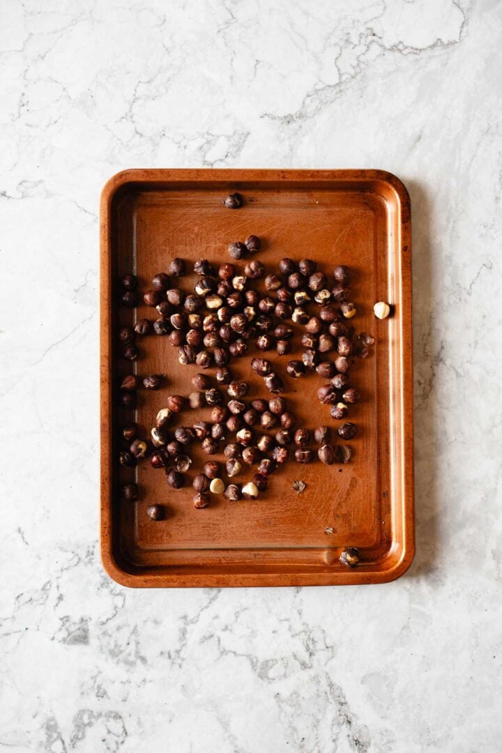 A wooden baking tray with scattered hazelnuts, some whole and some cracked, on a light marble surface.