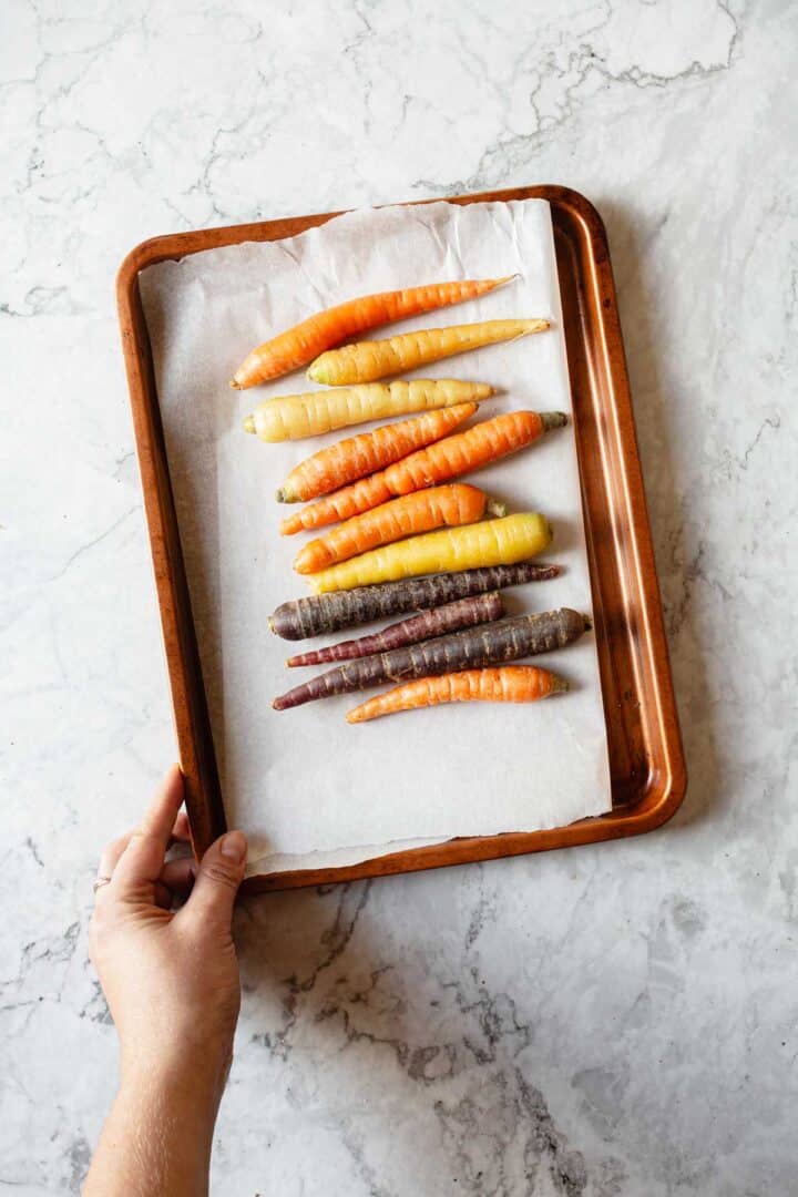 A hand holds a metal tray lined with parchment paper, displaying an assortment of colorful carrots, including orange, yellow, and purple varieties, arranged neatly on a marble countertop.