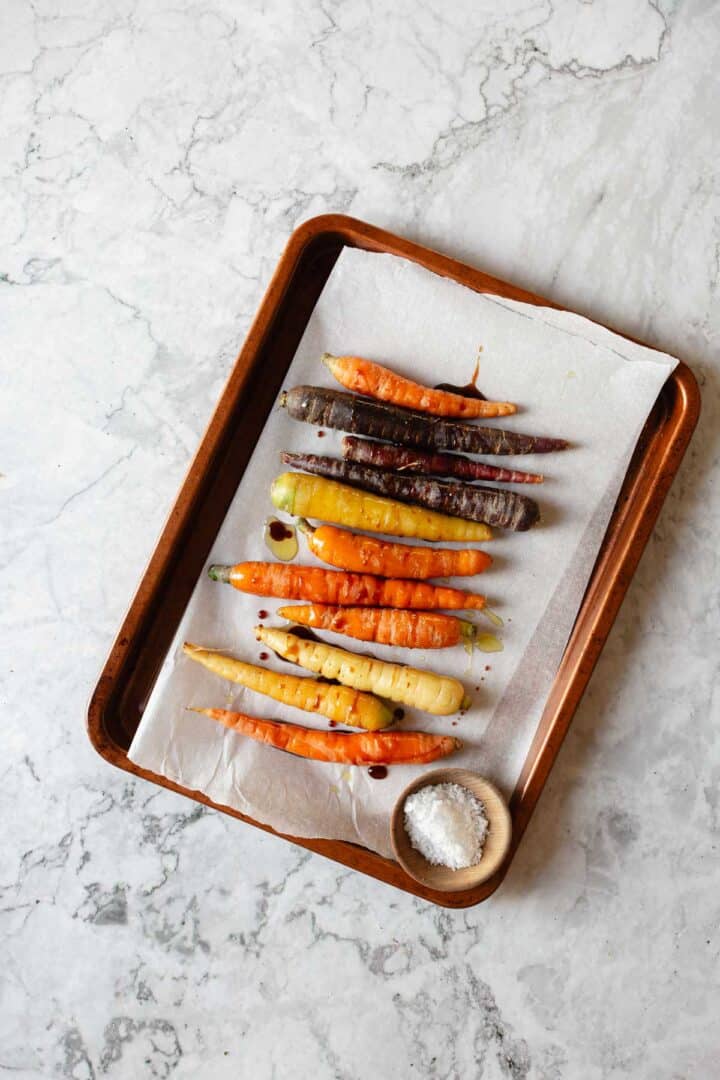 A baking tray with parchment paper displays a variety of colorful carrots, including orange, purple, and white. A small wooden bowl filled with coarse salt is placed beside the carrots on the tray.