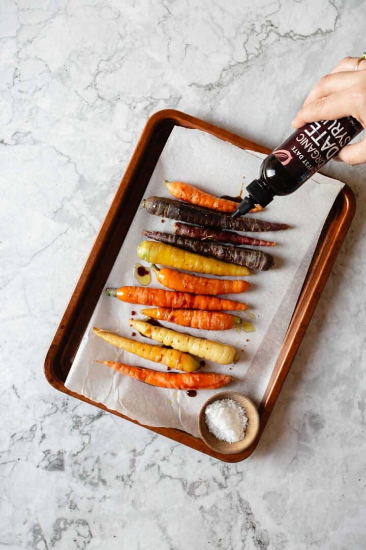 A baking tray with multicolored carrots on parchment paper is shown. A hand is drizzling balsamic glaze over the carrots. A small bowl of coarse salt is placed beside them on the tray. The background is a marbled surface.