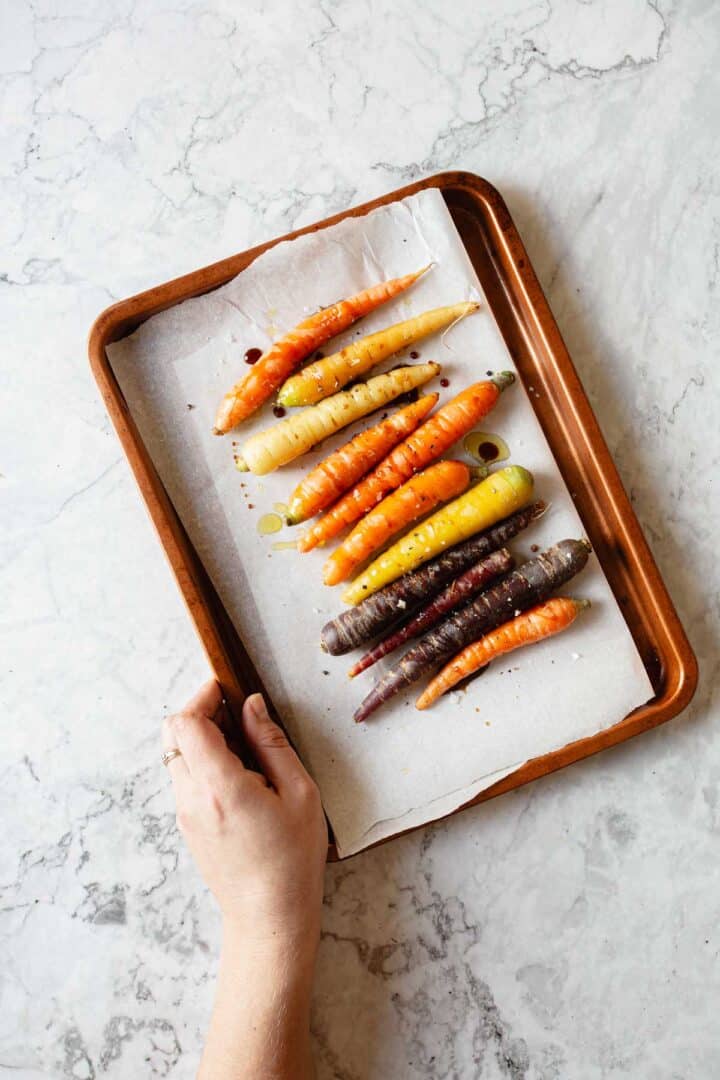 A hand holds a baking tray with assorted colorful carrots placed on parchment paper. The carrots are drizzled with oil and seasoning. The tray rests on a light marble surface.