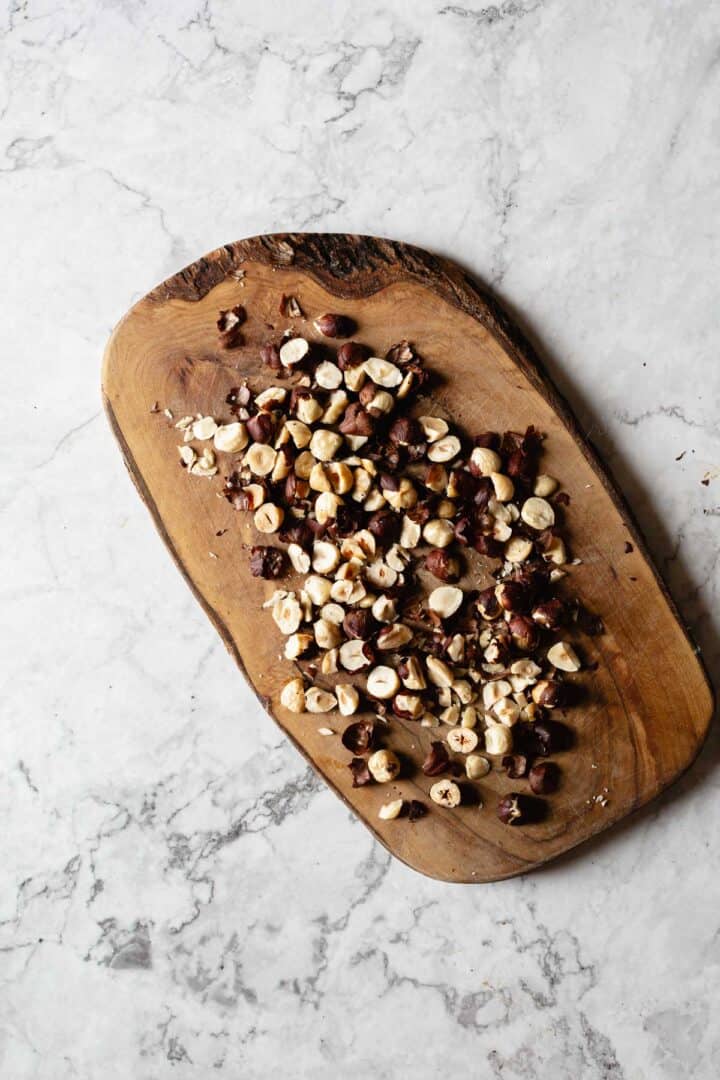 A wooden cutting board on a marble surface holds a pile of chopped hazelnuts.