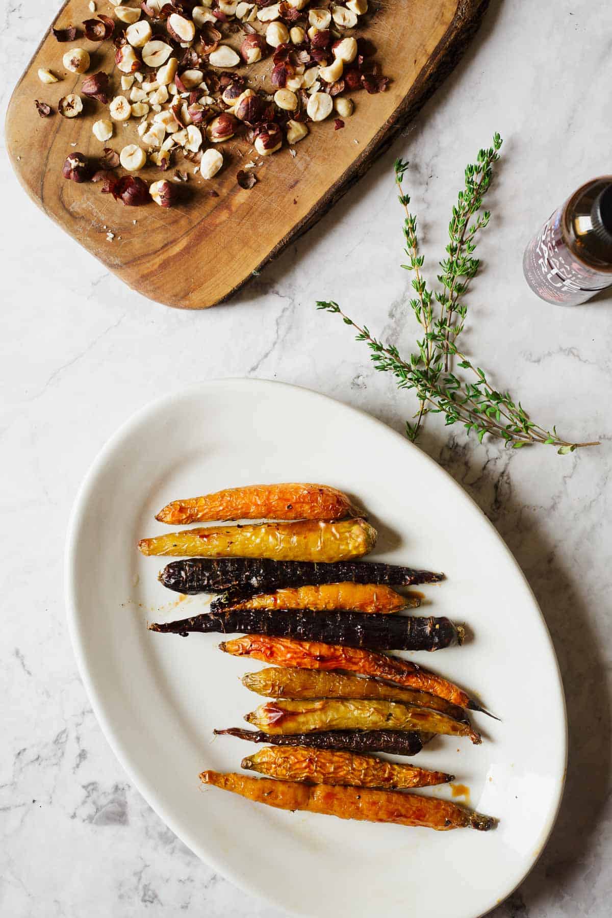 A white oval plate holds roasted multicolored carrots arranged in a row. Nearby, a wooden board displays chopped hazelnuts. Fresh thyme sprigs and a small bottle are also on the marble surface.