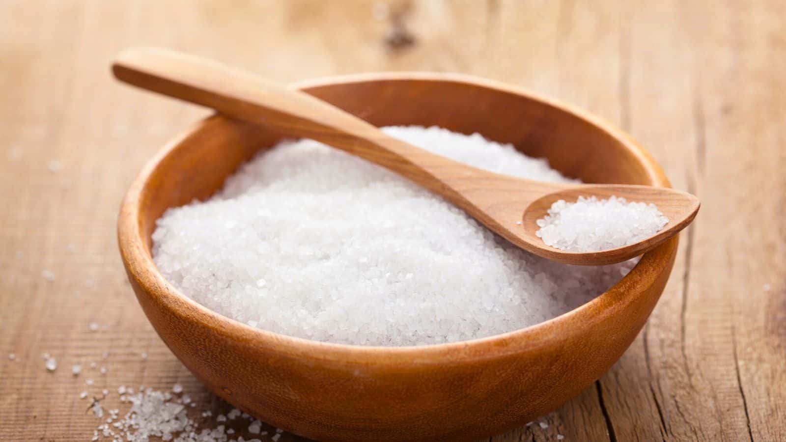 A wooden bowl filled with coarse-grained salt rests on a wooden surface. A small wooden spoon lies across the bowl, with a portion of salt heaped in its scoop. The background consists of a textured wooden tabletop.