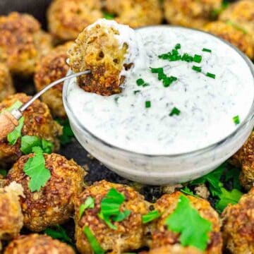 A plate of keto-friendly meatballs garnished with parsley surrounds a bowl of creamy sauce sprinkled with chopped chives. A fork holds one meatball, dipped in the delicious, low-carb sauce, above the bowl.
