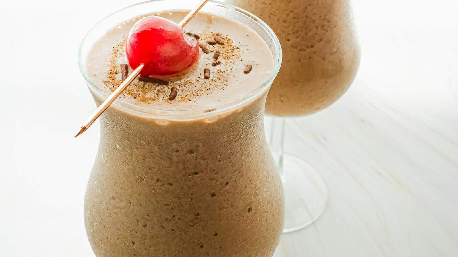 Two glasses of a frothy, brown beverage, likely a smoothie or milkshake, are shown on a light surface. Each drink is garnished with a maraschino cherry skewered on a toothpick and sprinkled with what appears to be chocolate shavings.