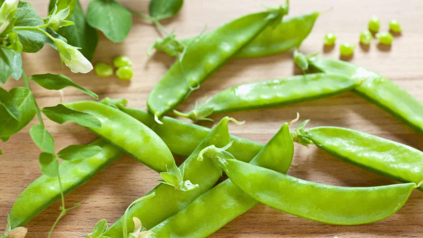 Fresh green snap peas with stems and leaves are scattered on a wooden surface. A few loose peas are visible in the background.
