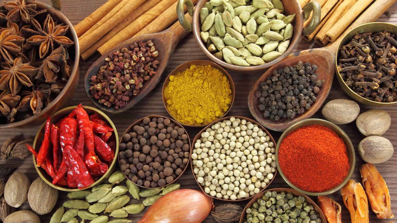 An assortment of spices displayed in wooden bowls and on a wooden surface, including cardamom, star anise, red chili peppers, black and white peppercorns, cloves, cinnamon sticks, nutmeg, mustard seeds, and allspice.