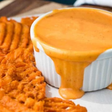 Waffle-cut sweet potato fries are arranged beside a white ramekin filled with creamy, orange cheese sauce, drizzling slightly down the side. Perfect game day snacks, this tasty arrangement is set on a wooden board with parchment paper.