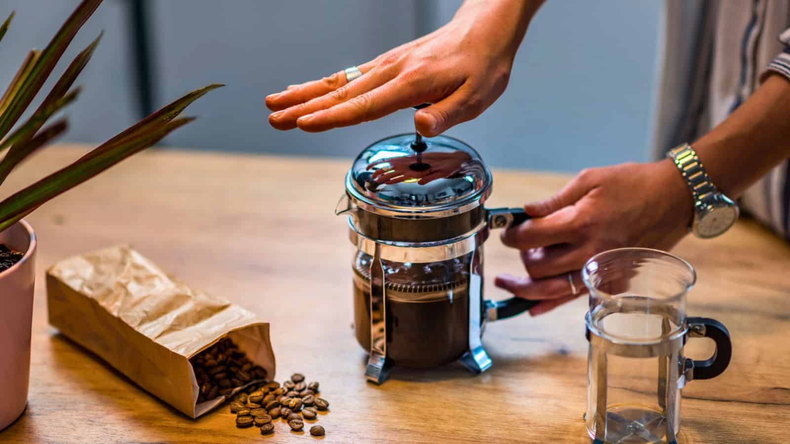 A person presses down the plunger on a French press filled with coffee grounds. The table holds a bag of coffee beans, scattered beans, and a glass of water. A potted plant is partially visible on the left.