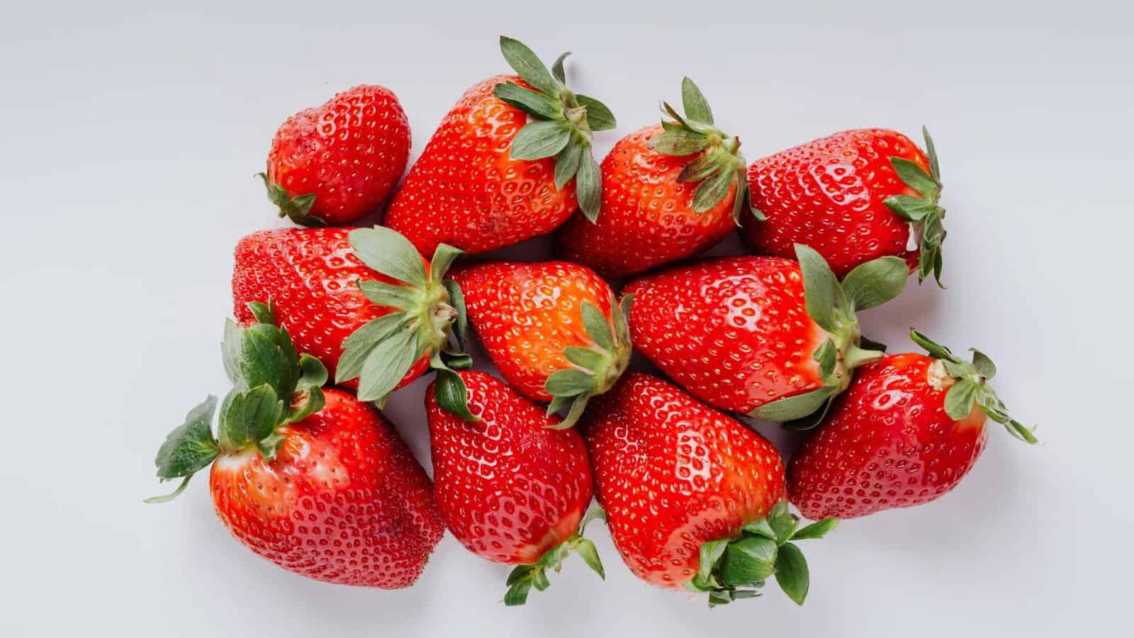 A cluster of ripe, red strawberries with green leaves is arranged on a plain white background. The strawberries vary slightly in size and shape, and their surfaces appear shiny and fresh.
