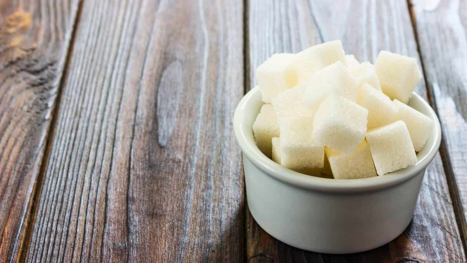 A white ceramic bowl filled with white sugar cubes sits on a wooden surface with visible grain patterns. The surface has a rustic appearance, and the focus is on the bowl, placed towards the right of the image.