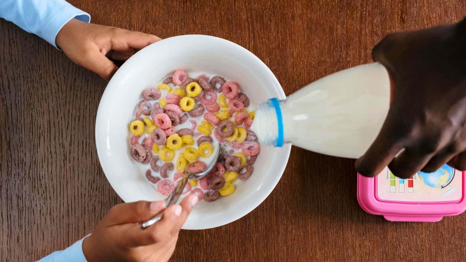 A person pours milk into a white bowl filled with colorful cereal loops. Another person holds a spoon, ready to eat. The bowl sits on a wooden table next to a pink container.