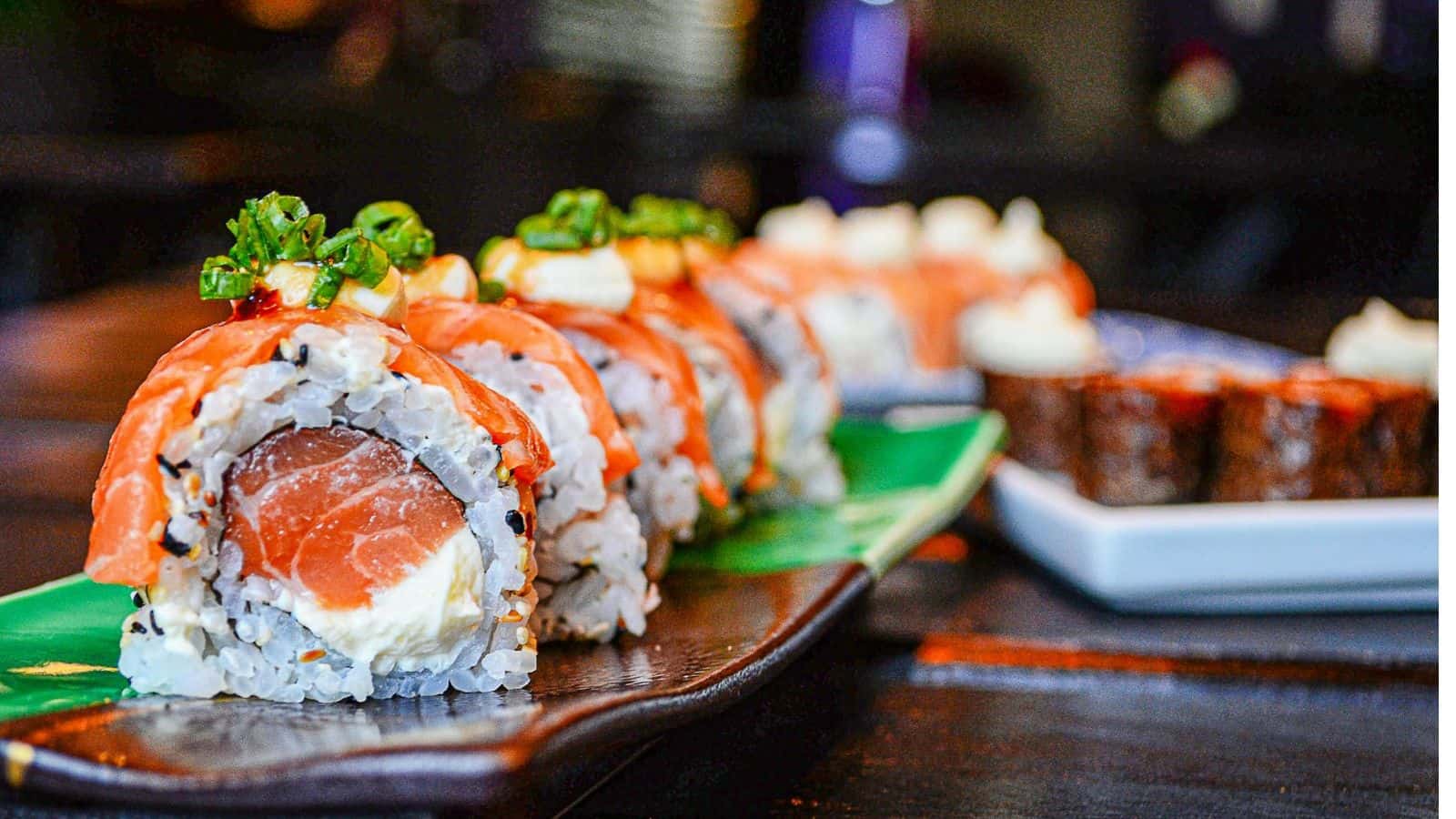 A close-up of sushi rolls arranged on a green plate. The rolls are topped with sliced salmon, cream cheese, and chopped green onions. A separate plate with more sushi rolls sits blurred in the background, on a dark wooden table.